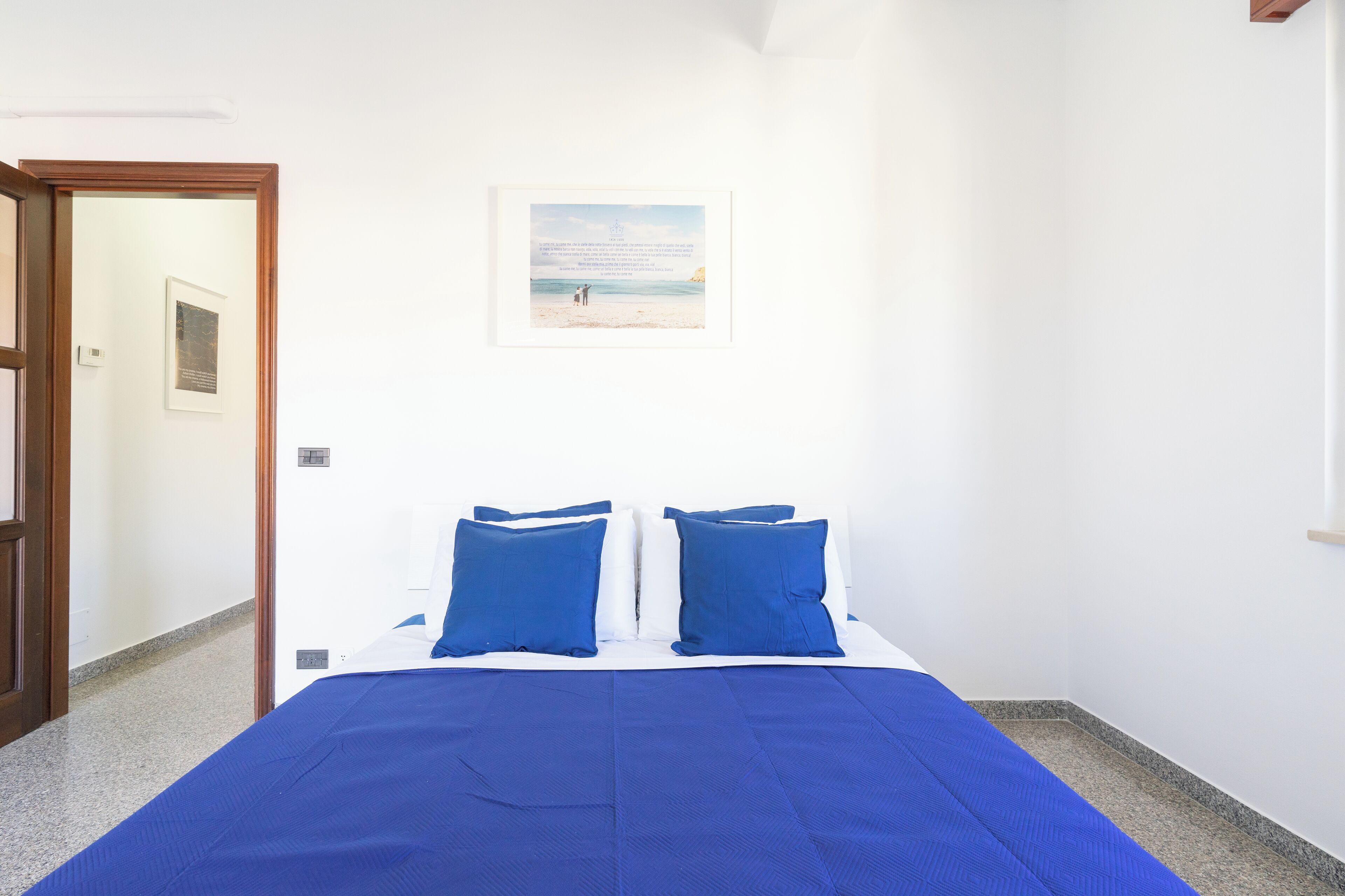 Une chambre à coucher avec un lit, deux oreillers bleus, un tableau encadré accroché au mur et une porte donnant sur une autre pièce.