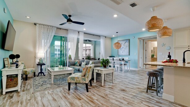 A spacious living room with a large window, a ceiling fan, and a dining area with a kitchen in the background.