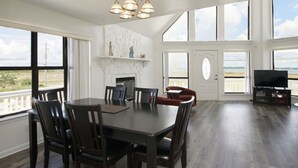 Ein geräumiger Essbereich mit großem Tisch und Stühlen, Kamin und Fernseher in einem Zimmer mit großen Fenstern und Blick auf den Strand.