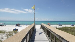 Ein Holzsteg führt zu einem Strand mit einer Fahnenmast und Strandsonnenschirmen.