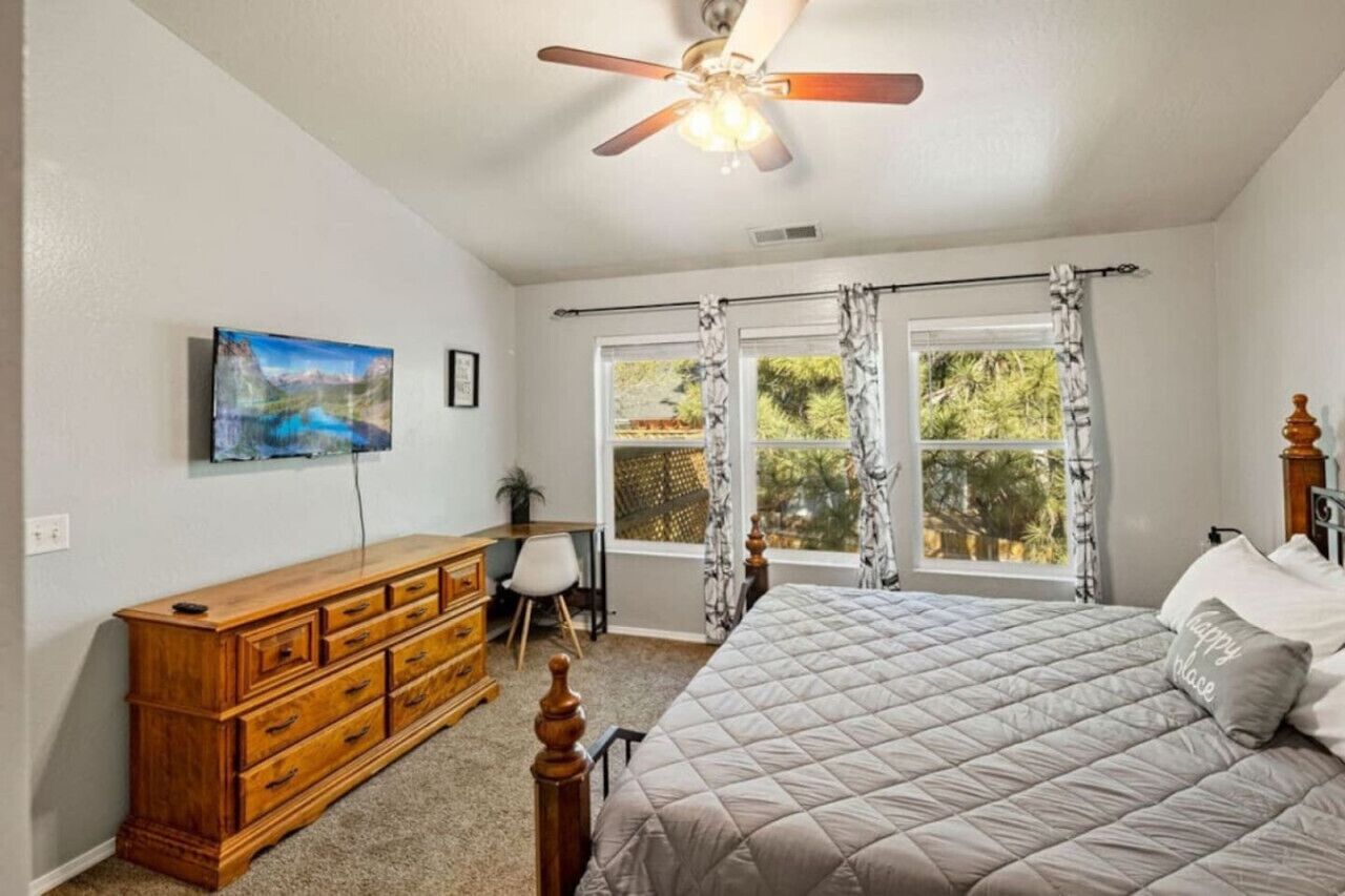 A bedroom with a bed, a wooden dresser, a desk, a chair, a ceiling fan, and a window with curtains.