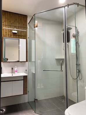 A modern bathroom with a glass-enclosed shower, a white sink with a mirror above it, and a wooden panel wall. - Lucy Hotel & Apartment (Ho Chi Minh City)