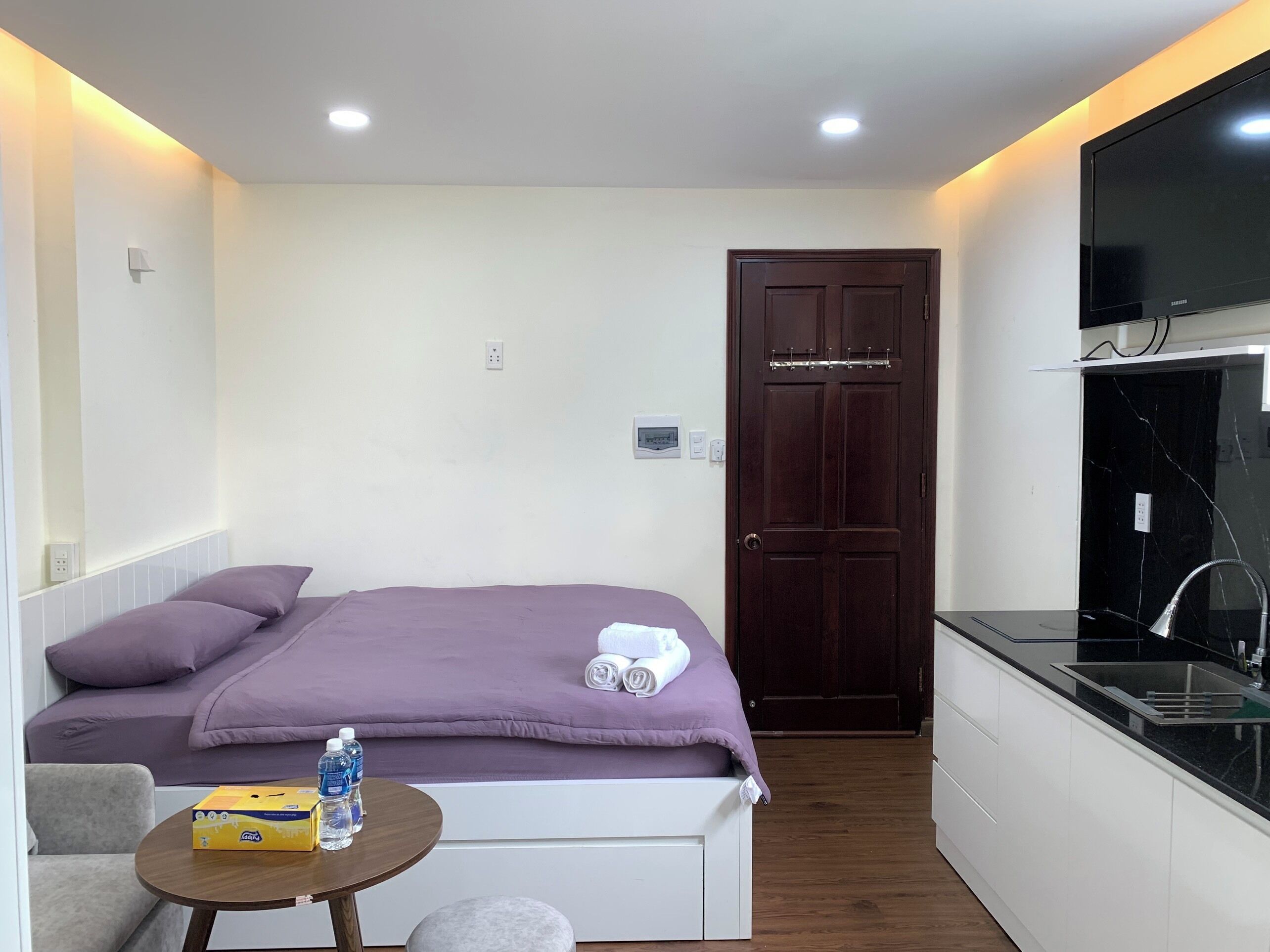 A compact room with a bed, a small table with water bottles, a sofa, a kitchenette, and a door.