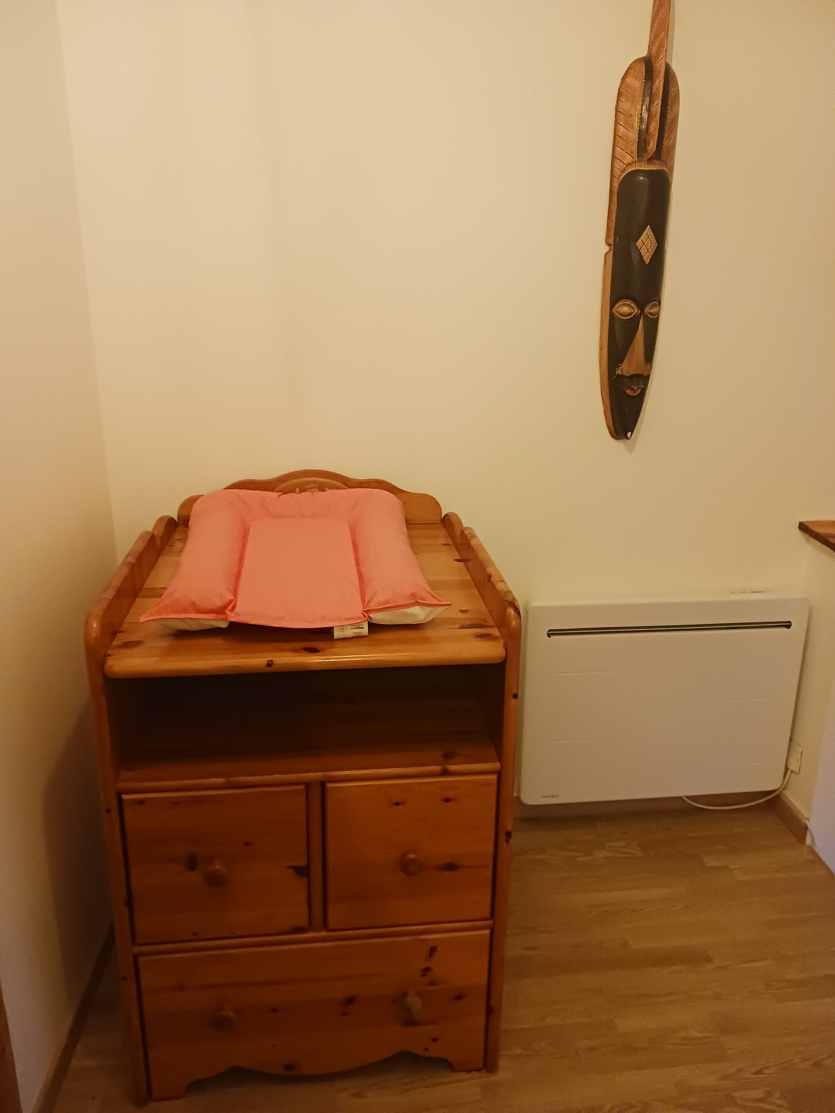 Eine Holzkommode mit rosa Bettwäsche darauf, eine Holzdekoration an der Wand und eine weiße Heizung.