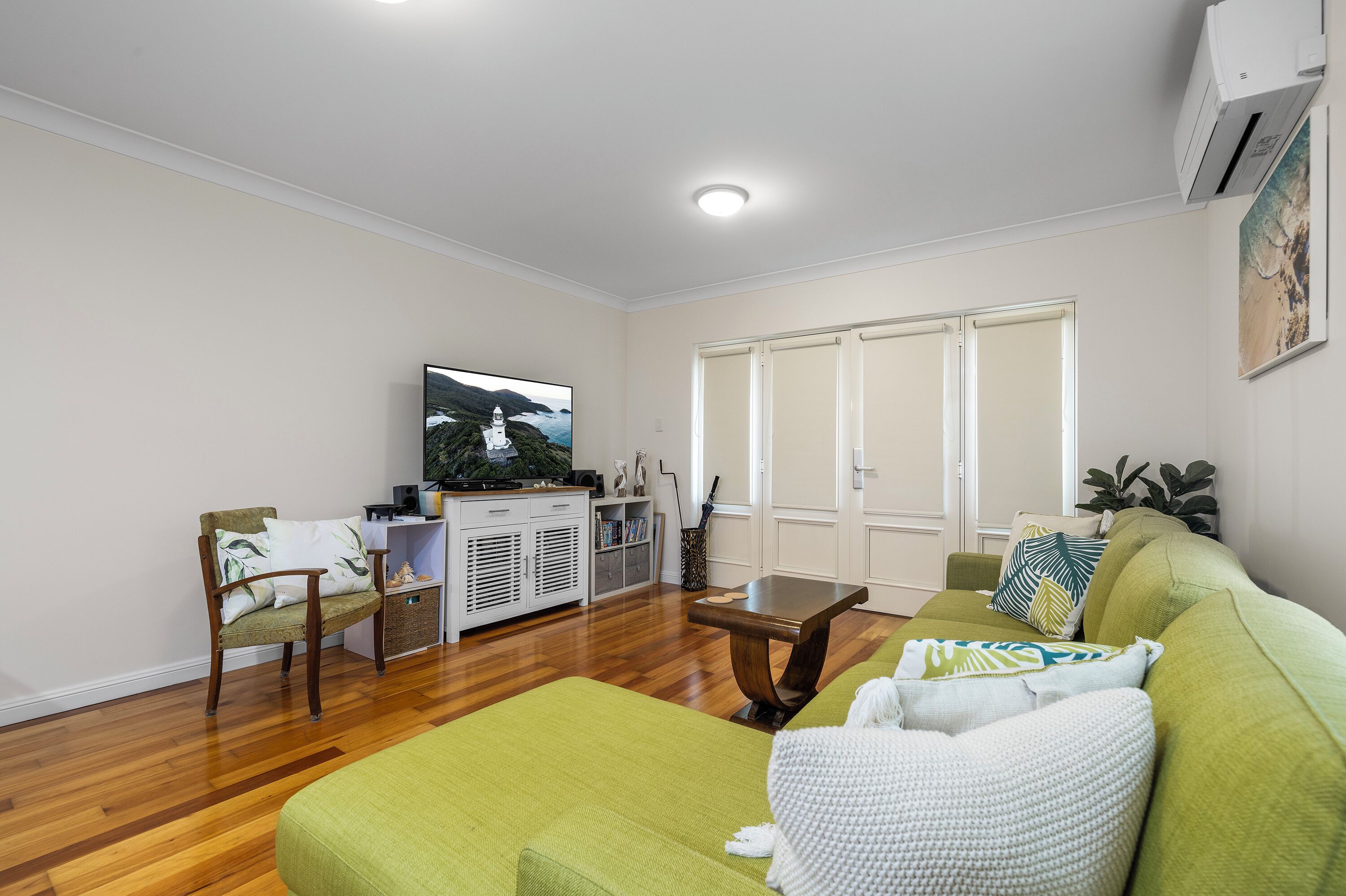 A living room with a green sofa, a wooden coffee table, and a television on a stand. There is a white cabinet with decorative items and a plant.