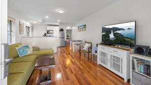A modern living room with a wooden floor, green sofa, dining table, and a television displaying a scenic view.