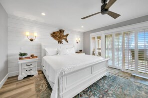 A bedroom with a large bed, a ceiling fan, a decorative wall piece, and a view through the window blinds. - Boardwalk 187 - Serene lake views with a boardwalk to the Gulf of Mexico! (Miramar Beach)