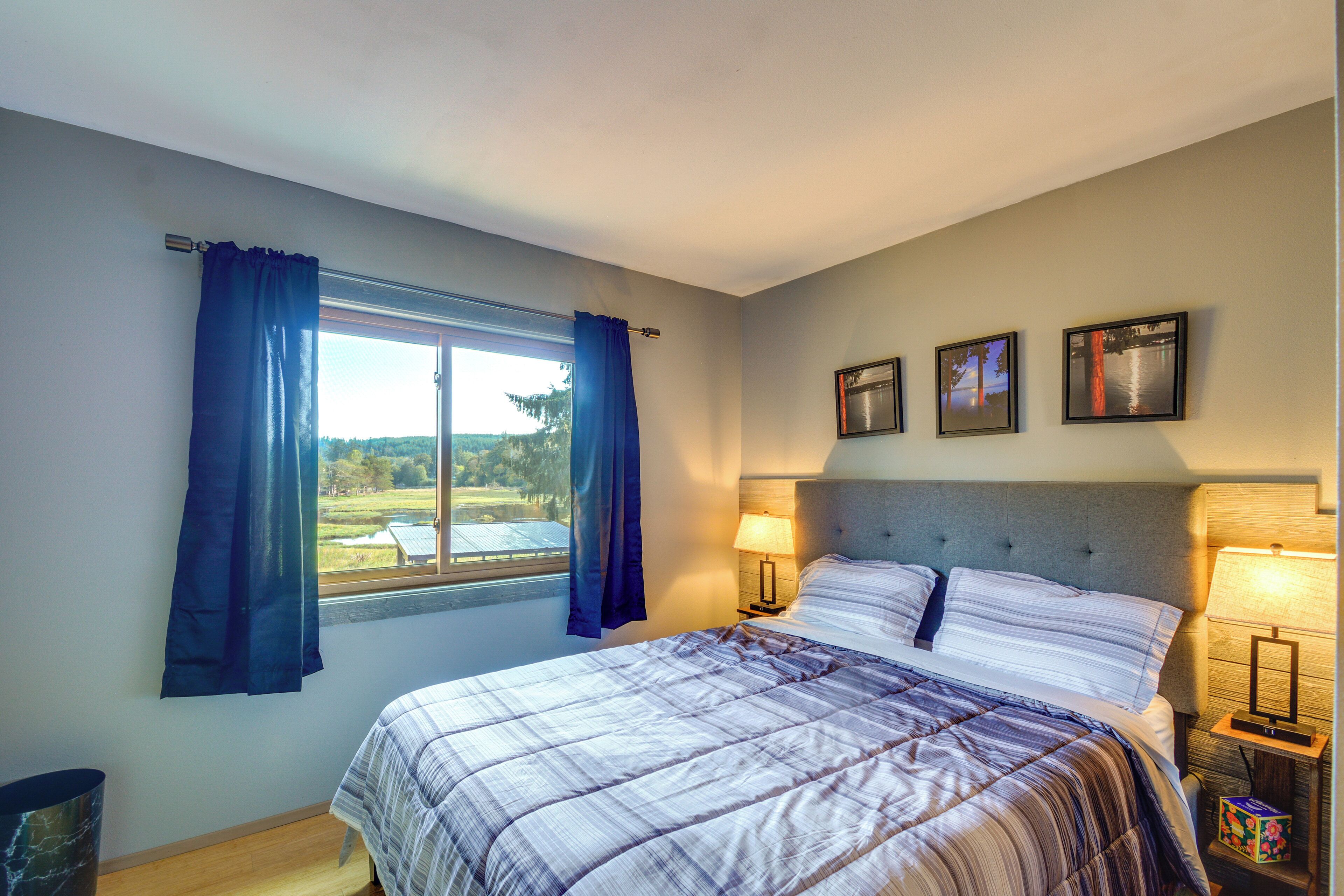 Une chambre à coucher avec un grand lit, une fenêtre avec des rideaux, une table de chevet avec une lampe et trois tableaux encadrés accrochés au mur.