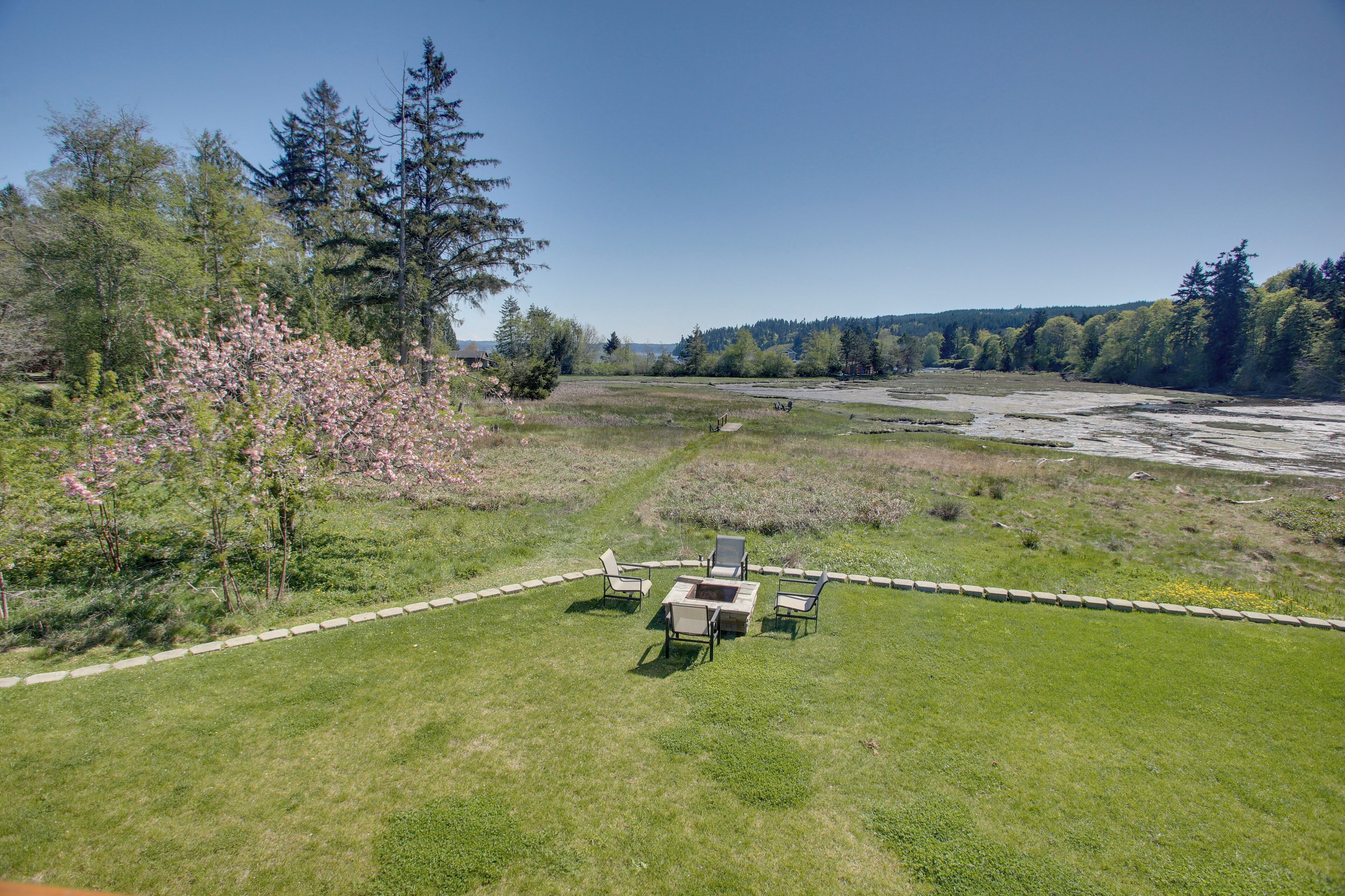 Un espace extérieur verdoyant, doté d’un foyer et de sièges, entouré d’arbres et offrant une vue sur une rivière.