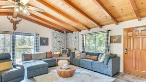 Un salon chaleureux avec une cheminée en pierre, des poutres apparentes en bois au plafond et une grande fenêtre donnant sur une forêt.