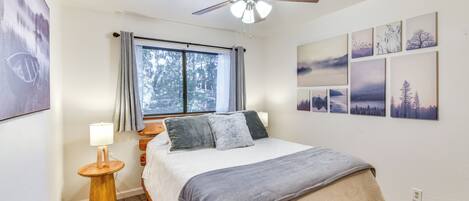 Un dormitorio con cama, ventilador de techo, ventana con cortinas, mesita de noche con lámpara y pared decorada con cuadros.