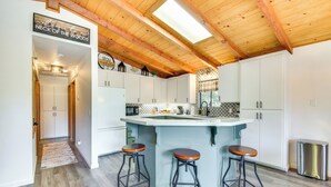 Une cuisine avec un plafond en bois, des armoires blanches, un îlot central avec des tabourets et une pancarte sur laquelle on peut lire : « Bienvenue à Neck of the Woods ».