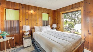 A bedroom with wooden walls, a large bed with white bedding, two bedside tables with lamps, and a window with a view of a house and trees. - Waterfront Cottage w/ Dock & Views in Six Lakes! (Six Lakes)