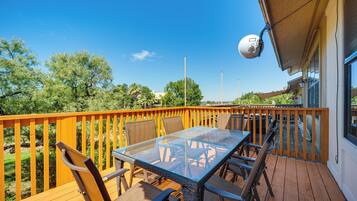 A deck with a glass table and chairs, surrounded by a wooden railing, overlooking a landscaped area with trees and a clear sky.