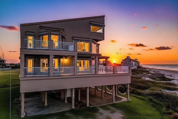 A two-story beachfront house with multiple balconies and a view of the sunset over the ocean.