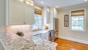 Una cocina con armarios blancos, encimeras de granito, un refrigerador de acero inoxidable y una ventana con persiana de bambú.