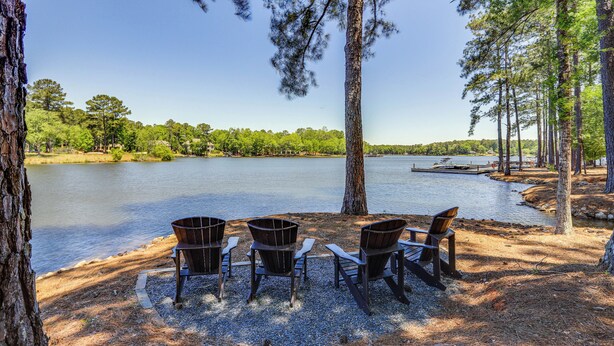 Zona de estar junto al lago con tres sillas Adirondack y un banco de madera, rodeada de árboles y una vista despejada del lago.