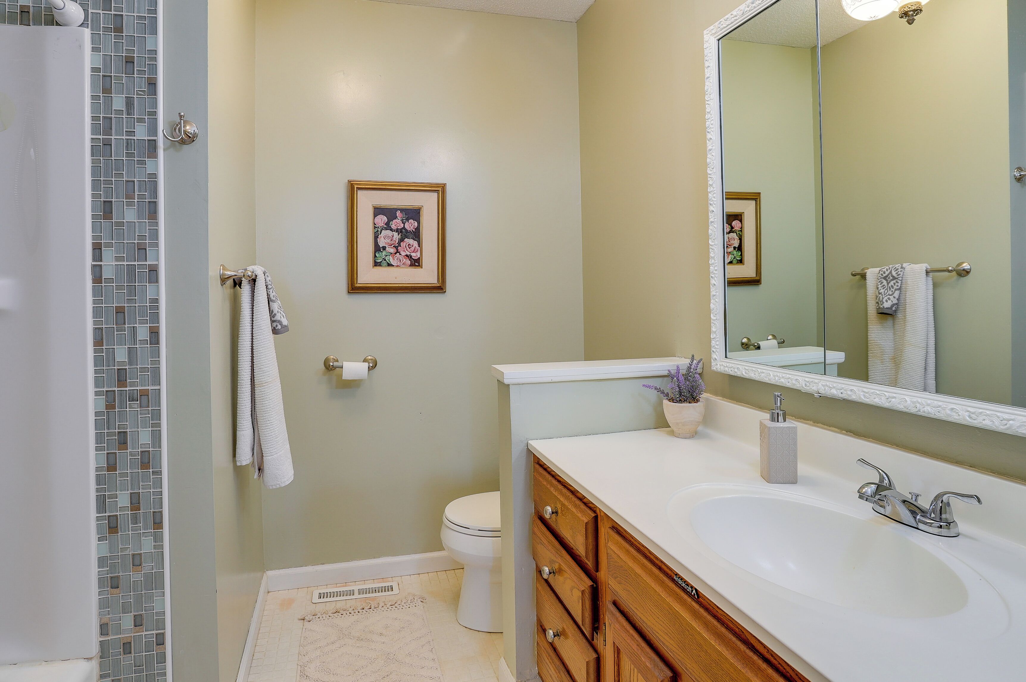 A bathroom with a white sink, wooden vanity, a large mirror, a framed picture on the wall, and a shower area with a tiled wall.