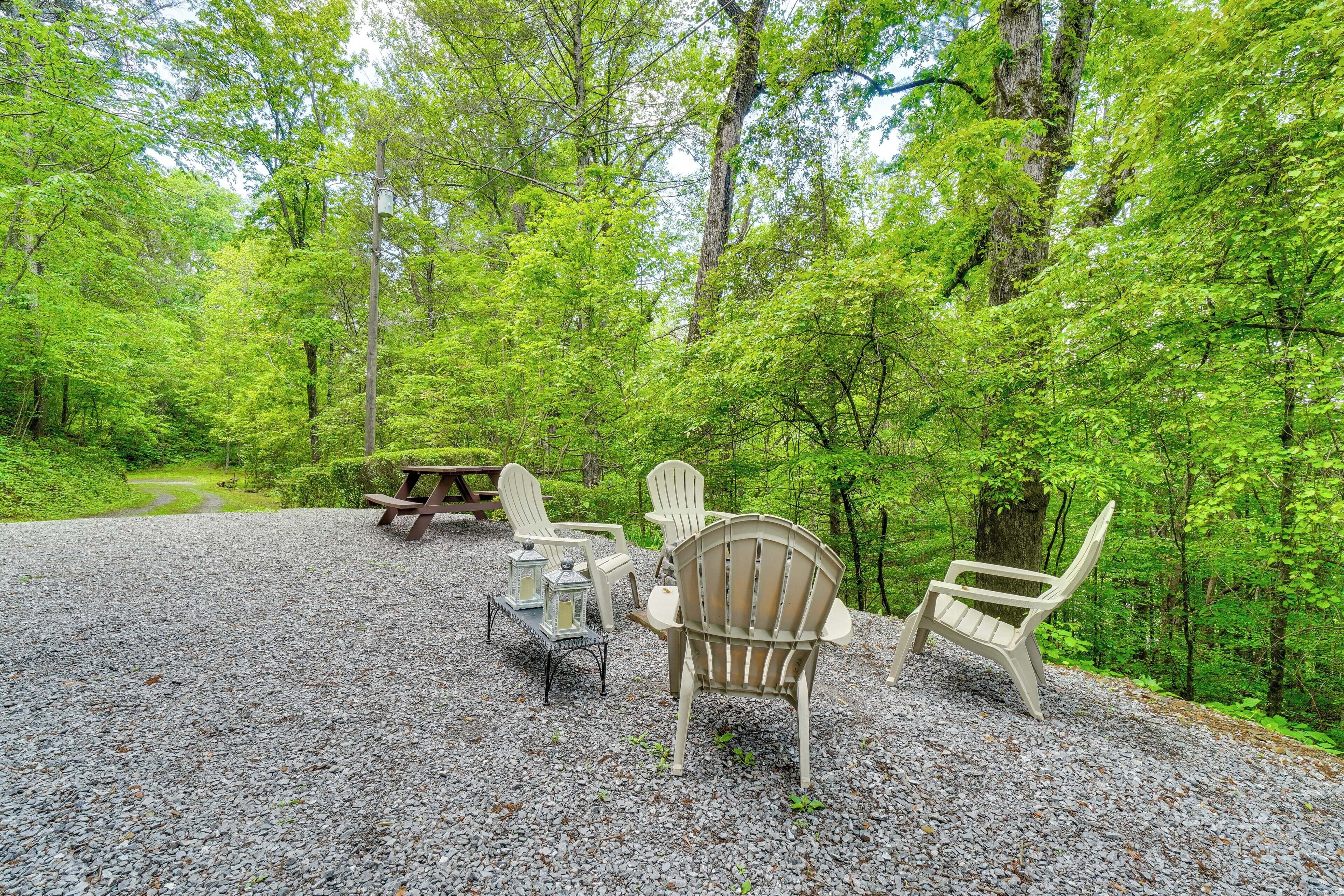 Un espacio con grava, sillas Adirondack y una mesa de picnic, rodeado de un bosque frondoso.