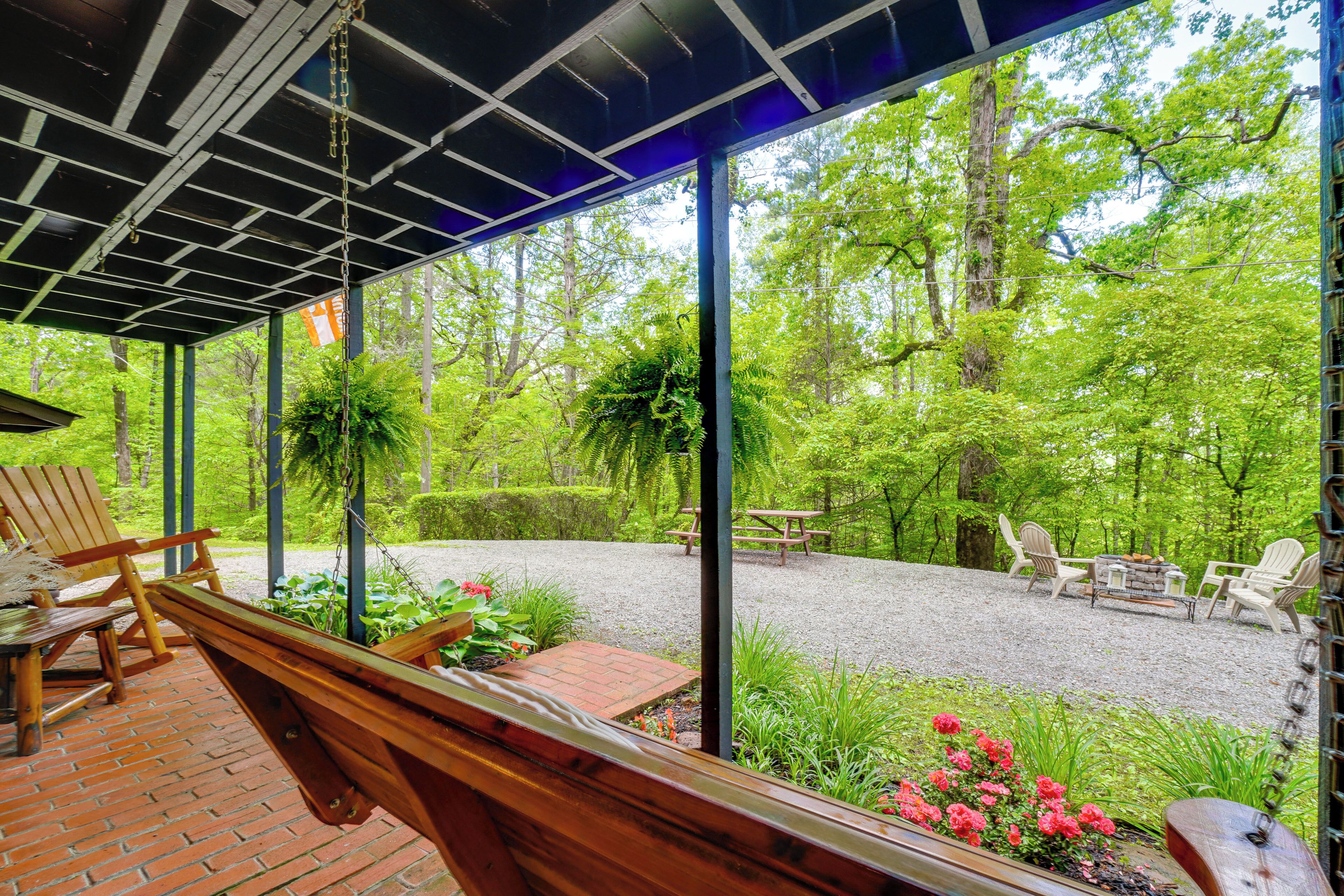 A covered outdoor seating area with wooden benches and a picnic table, surrounded by lush greenery and trees.