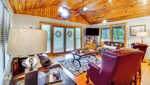 A spacious living room with a wooden ceiling, a ceiling fan, a fireplace, a television, and a large window with blinds. - 'bear Acres Getaway' Cabin w/ Deck in Townsend! (Townsend)