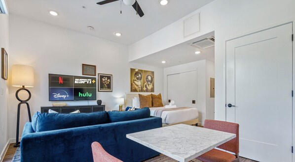 A modern hotel room with a blue sofa, a white bed, a marble coffee table, and a TV displaying streaming services logos. - Vibrant Austin Studio | Balcony + Parking + Wi-Fi (Austin)