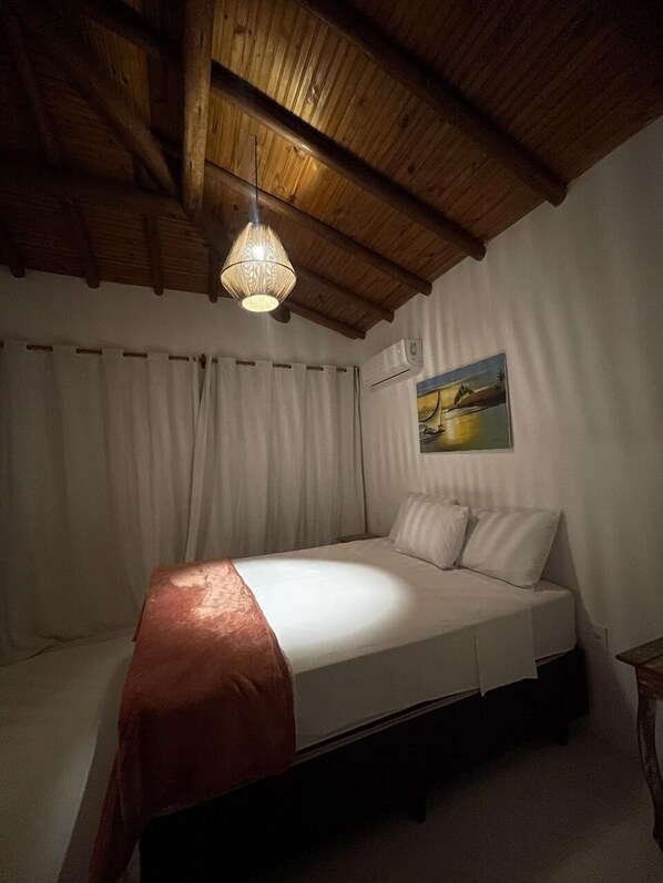 A bedroom with a wooden ceiling, a bed with a white mattress and pillows, a hanging light fixture, and a painting on the wall.