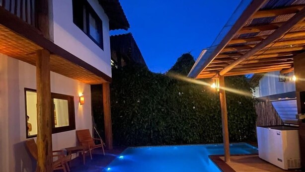 A swimming pool with wooden deck chairs, a covered patio area, and a white building with a balcony. - Casa nobre Trancoso  (Porto Seguro)