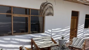 An outdoor seating area with wooden furniture, a woven hanging light fixture, and a view of the sea through a large window. - HOP MAR Surfea las Mejores Olas de Puemape (Puémape)