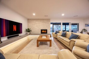 A modern living room with a large flat-screen TV, a stone fireplace, a beige sofa, and a wooden coffee table. - Galene Getaway Walk to Beach Pool Fire (Mount Martha)
