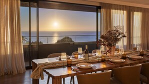 Een eetruimte met een tafel voor zes personen, grote ramen met uitzicht op zee en een potplant op tafel.