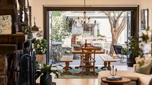 A cozy living room with a fireplace, a dining area with a table and chairs, and a view of the outdoors. - Bella s Cottage Spa Sauna Large Groups (Yarra Glen)