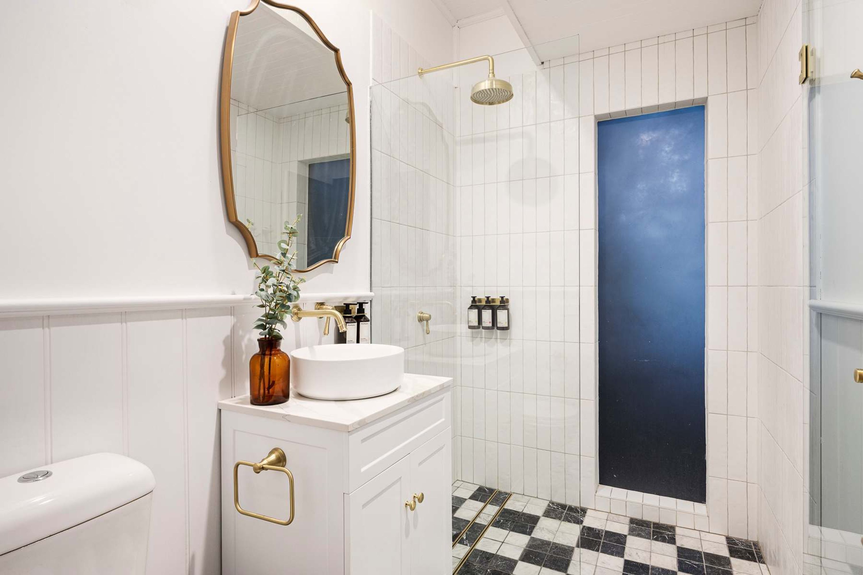Un baño con tocador blanco, espejo con marco dorado, inodoro blanco y ducha con puerta azul.