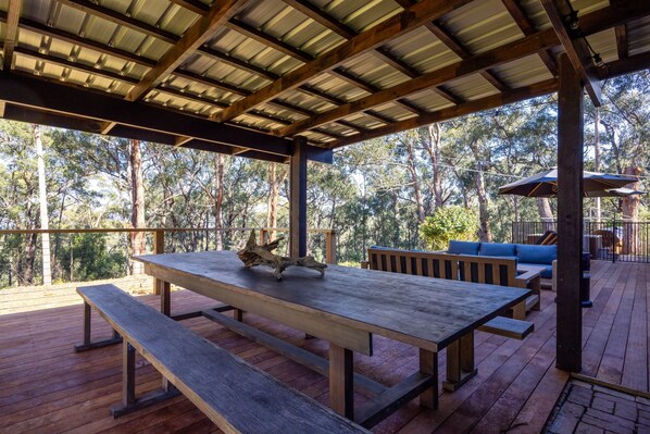 Una terraza de madera al aire libre con una mesa de picnic y bancos, rodeada de árboles.