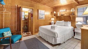 Habitación de hotel con detalles de madera, una cama con ropa blanca, una silla azul y un pequeño escritorio con una lámpara.