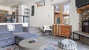 Un salon moderne avec un canapé d’angle gris, une table basse ronde et un tapis à motifs. On y trouve un meuble en bois, un mur d’accent en pierre et un téléviseur fixé au-dessus.
