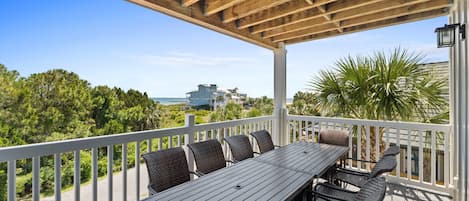 A covered outdoor deck with a dining table and chairs, overlooking a scenic view of a beach and ocean.