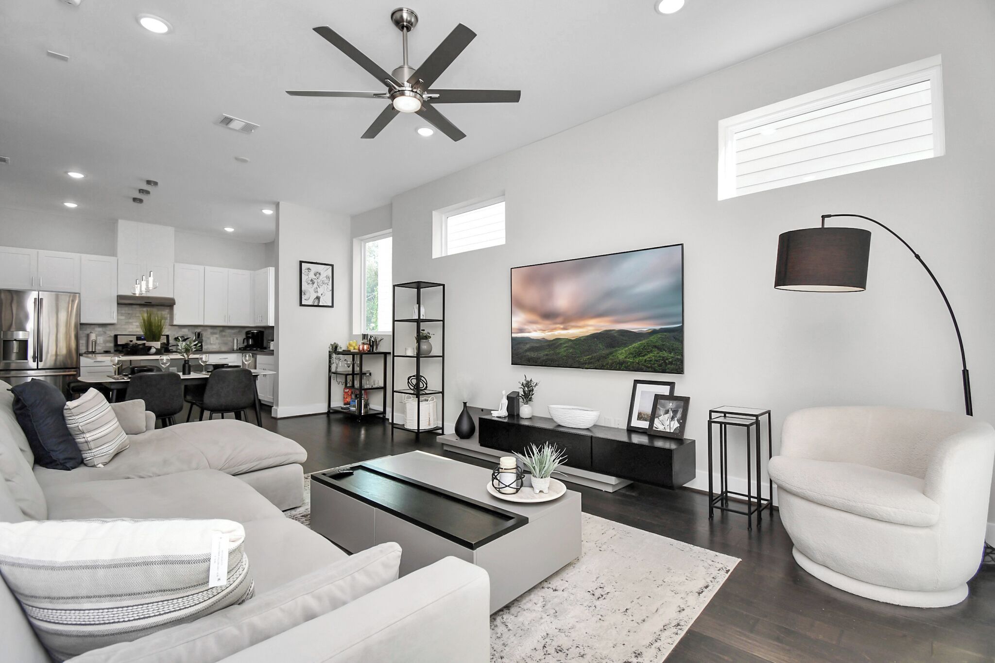 A modern living room with a sofa, coffee table, and a TV. There is a kitchen in the background with cabinets and a dining area.