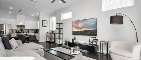 A modern living room with a sofa, coffee table, and a TV. There is a kitchen in the background with cabinets and a dining area.