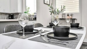 Un espace repas moderne avec une table blanche dressée pour deux personnes, agrémentée de bols noirs et de verres à vin transparents.