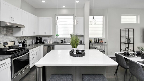 Une cuisine moderne avec des meubles blancs, un îlot central, des appareils en acier inoxydable et des luminaires suspendus.