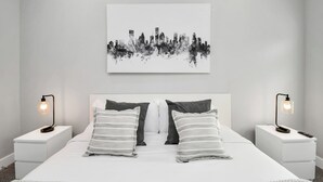Une chambre à coucher moderne, ornée d’une œuvre d’art représentant un paysage urbain, avec deux tables de chevet équipées de lampes et un lit agrémenté d’oreillers à rayures.
