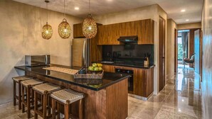 A modern kitchen with wooden cabinets, a central island with stools, hanging pendant lights, and a view to the outside. - 3BR Deluxe PH w Private Pool + Rooftop (Tulum)