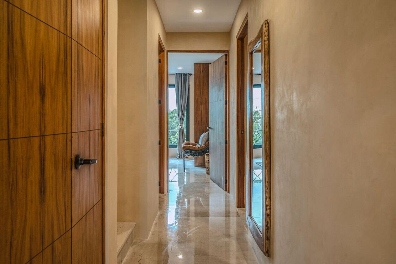 A hallway with wooden doors and a view into a room with a chair and a window.