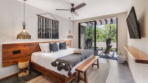 Une chambre à coucher moderne avec une tête de lit en bois, un grand lit agrémenté de plusieurs oreillers, un banc, un ventilateur de plafond et une vue sur la verdure à travers la porte vitrée.