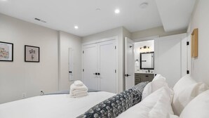 A modern bedroom with a neatly made bed, white walls, and a large mirror above the vanity.