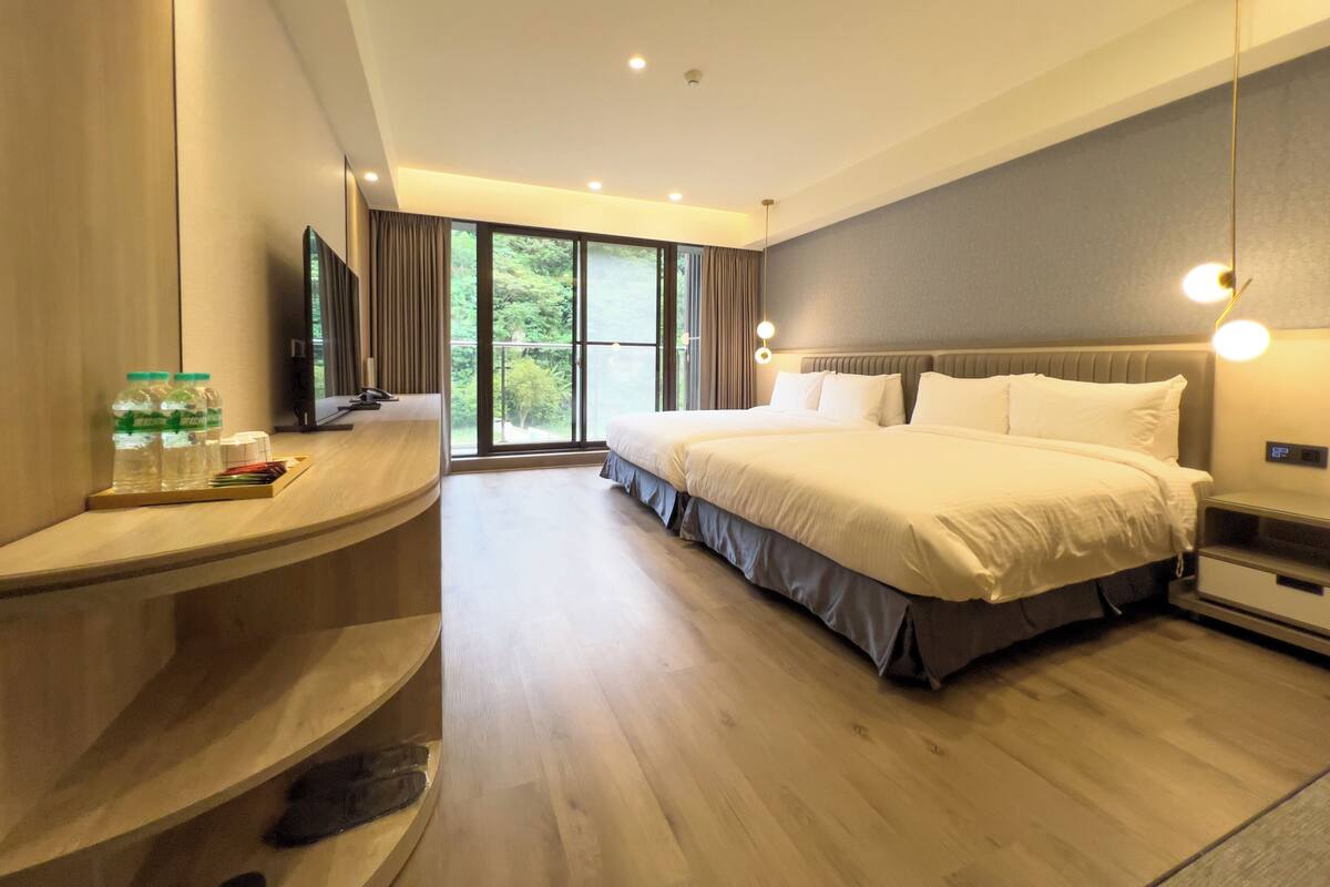 Une chambre d’hôtel moderne, dotée d’un grand lit, d’un bureau avec un ordinateur et offrant une vue sur la verdure par les fenêtres.
