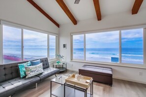 A spacious living room with a large window overlooking the ocean, a gray sofa with decorative pillows, a glass coffee table, and a brown bench.