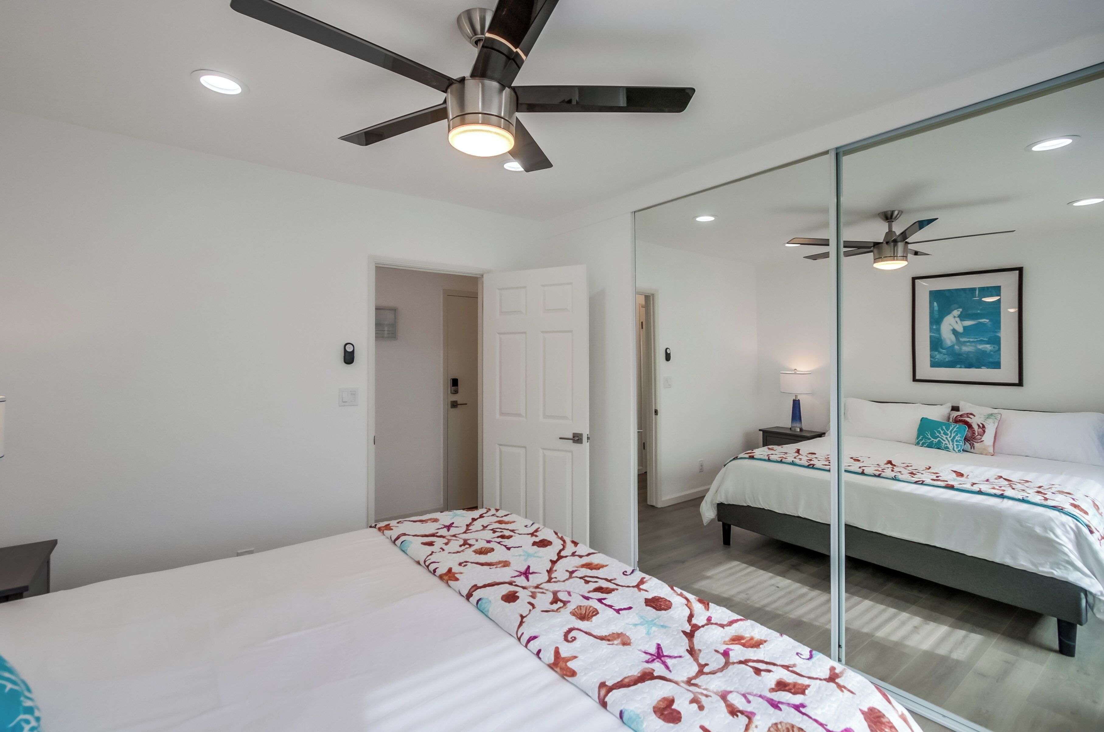 A hotel room with a bed, a ceiling fan, a mirror, and a painting on the wall.