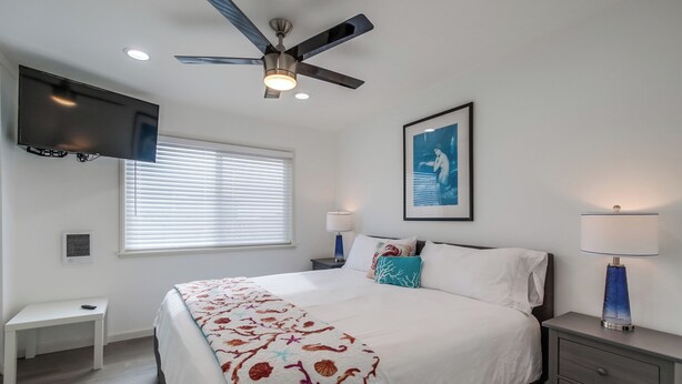 A bedroom with a bed, a ceiling fan, a TV mounted on the wall, a window with blinds, a bedside table, a nightstand with a lamp, and a framed picture on the wall. - Right on the Famous Mission Beach Boardwalk - Sleeps up to 10 - Best location in Mission Beach (San Diego)